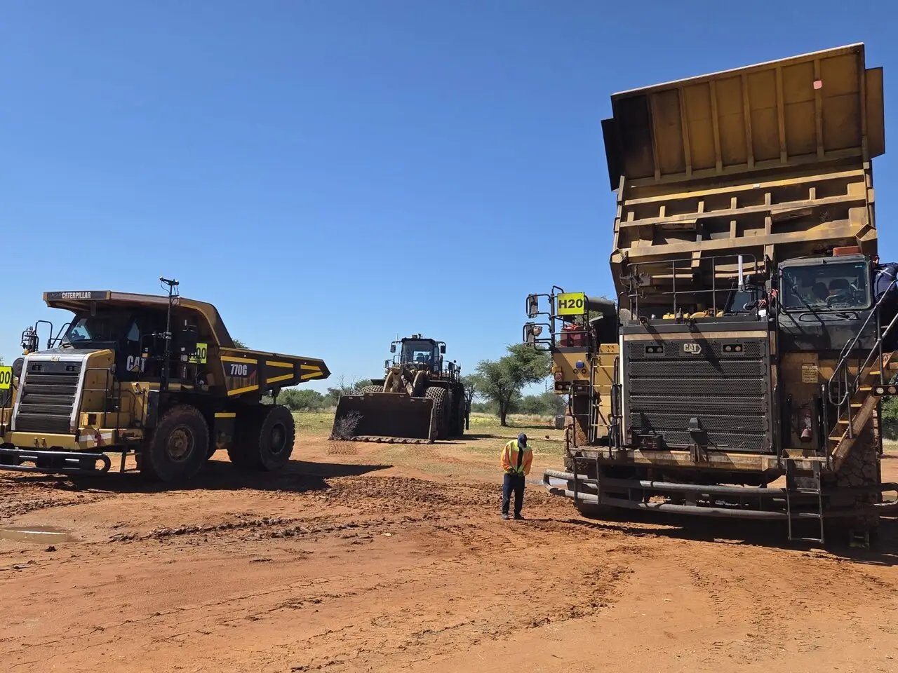 Haul trucks and a front-end loader parked at a mine site with a person in high-visibility clothing